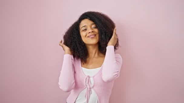 Mujer afroamericana sonriendo con confianza peinando el cabello con las manos sobre el fondo rosa aislado - Metraje, vídeo