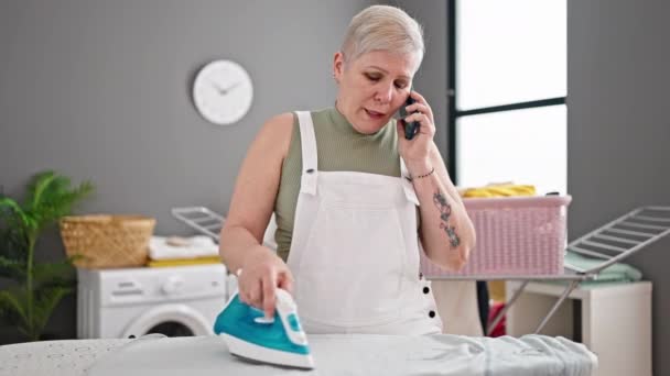 洗濯室でスマートフォンのアイロン掛け服で話す中年の白髪の女性 - 映像、動画