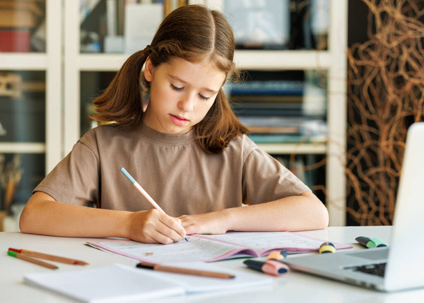 Koncentrált gyermek alkalmi ruhák ül az asztalnál, és írás tervező közelében laptop alatt online lecke napközben otthon - Fotó, kép
