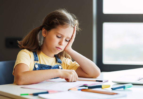 frustrovaná dívka přemýšlí nad obtížným úkolem a dotýká se hlavy, zatímco dělá domácí úkol u stolu - Fotografie, Obrázek