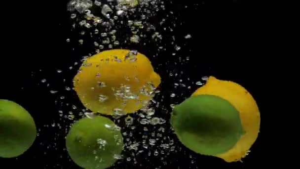 Медленно движутся зеленая липа и желтый лемон, падающие в прозрачную воду на черном фоне. Брызги свежих фруктов в аквариуме. Цитрусовые, продукты питания, пузырьки воздуха - Кадры, видео