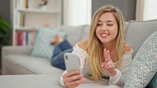 Giovane donna bionda scattare foto selfie con smartphone sdraiato sul divano a casa - Filmati, video