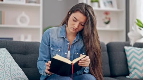 Νεαρή όμορφη ισπανόφωνη γυναίκα διαβάζει το βιβλίο κάθεται στον καναπέ στο σπίτι - Πλάνα, βίντεο