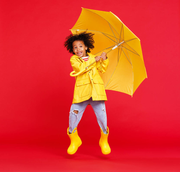 Lustiger afrikanisch-amerikanischer Junge in Regenmantel und Stiefeln mit Regenschirm springt, während er an Regentagen vor rotem Hintergrund läuft - Foto, Bild