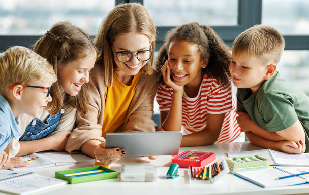 Tutor feminino feliz e crianças otimistas sorrindo e assistindo vídeo no tablet enquanto se reúnem em torno da mesa durante a aula na escola - Foto, Imagem