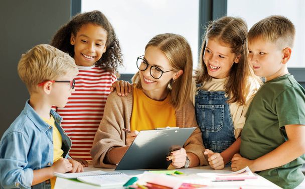 Счастливая воспитательница и оптимистичные дети улыбаются и обсуждают тему урока, собирая за школьным столом - Фото, изображение