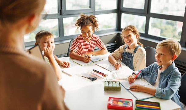 Γυναίκα δάσκαλος διεξάγει ένα μάθημα και εξηγεί ένα νέο θέμα σε αισιόδοξα παιδιά, ενώ μαζεύονται γύρω από το τραπέζι σε μια φωτεινή τάξη στο σχολείο - Φωτογραφία, εικόνα