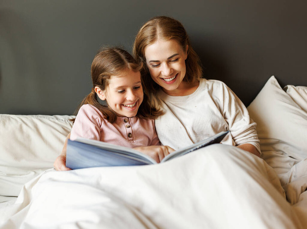 Wesoła rodzina: kobieta i dziewczyna uśmiechnięte i czytające bajki podczas relaksu na łóżku w pobliżu lampy w domu - Zdjęcie, obraz
