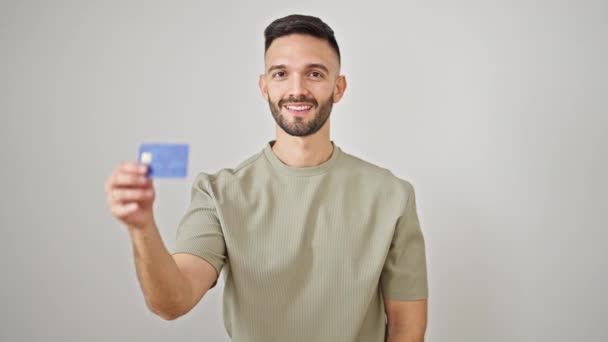 Joven hombre hispano sonriendo confiado sosteniendo tarjeta de crédito sobre fondo blanco aislado - Imágenes, Vídeo