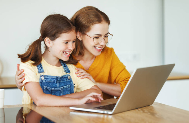 Chica alegre con su madre usando un ordenador portátil o haciendo clases en línea en la cocina en casa - Foto, imagen