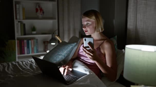 Mujer rubia joven usando teléfono inteligente y portátil sentado en la cama en el dormitorio - Metraje, vídeo