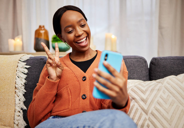Selfie, donna nera e segno di pace a casa con un sorriso per i social media, motivazione e videochiamata. Foto, persona femminile felice e africana in un soggiorno con un gesto della mano v ed emoji a casa. - Foto, immagini