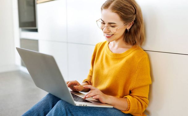 cheerful young woman uses a laptop at home sitting on the floor in a modern kitchen - Photo, Image