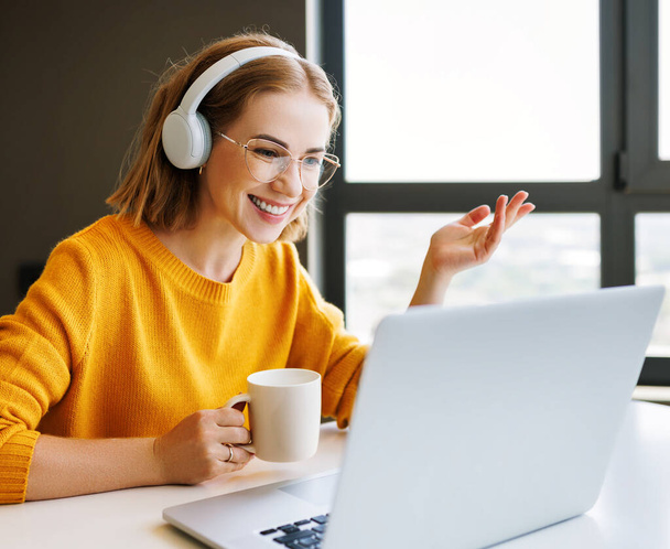 Fröhlich fröhliche junge Unternehmerin mit Brille lächelt und gestikuliert, während sie am Schreibtisch im Home Office sitzt und während eines Online-Meetings mit Online-Mitarbeitern spricht - Foto, Bild