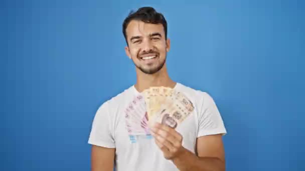 Genç İspanyol adam gülümsüyor. Meksika pesolarını soyutlanmış mavi arka planda tutuyor. - Video, Çekim