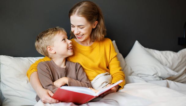 Cheerful family: woman and boy son smiling and reading fairy tale while relaxing on bed  at home - Photo, Image