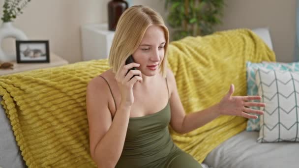 Mujer rubia joven hablando por teléfono sentado en el sofá discutiendo en casa - Imágenes, Vídeo