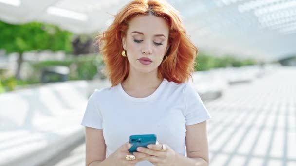 fiatal vörös hajú nő használ smartphone mosolygós parkban - Felvétel, videó