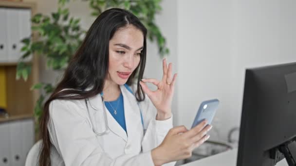 Young beautiful hispanic woman doctor doing video call with smartphone at the clinic - Footage, Video