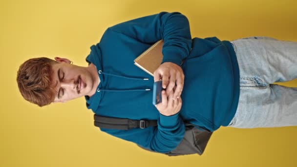Fiatal spanyol férfi diák okostelefon olvasás könyv mosolygós izolált sárga háttér - Felvétel, videó