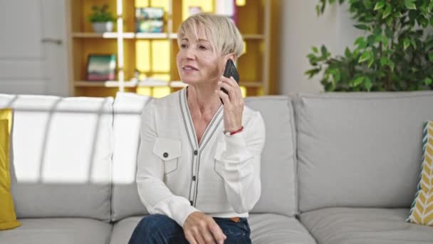 Középkorú szőke nő beszél telefonon ül a kanapén otthon - Felvétel, videó
