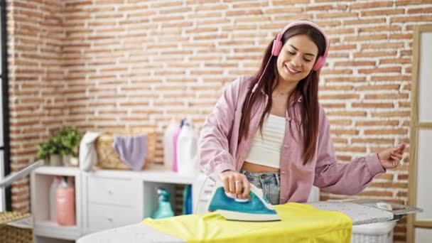Joven hermosa mujer hispana escuchando música planchando ropa bailando en la lavandería - Imágenes, Vídeo
