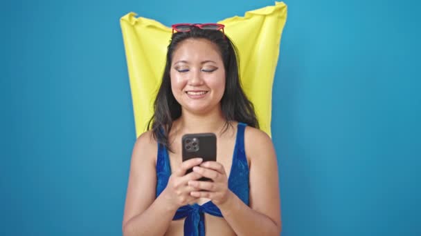 Junge chinesische Touristin trägt Bikini mit Smartphone über Schwebematratze vor isoliertem blauen Hintergrund - Filmmaterial, Video