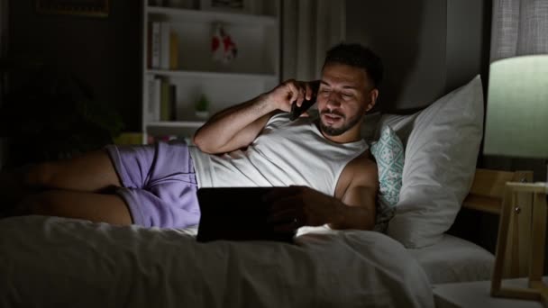 Hombre árabe joven hablando en el teléfono inteligente utilizando touchpad acostado en la cama en el dormitorio - Imágenes, Vídeo