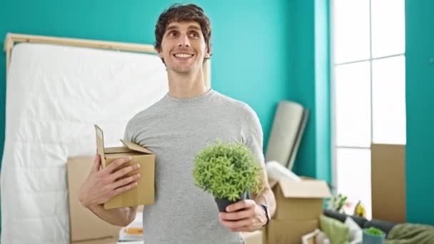 Genç İspanyol adam gülümsüyor. Yeni evinde kendine güvenen bir paket ve bitki tutuyor. - Video, Çekim