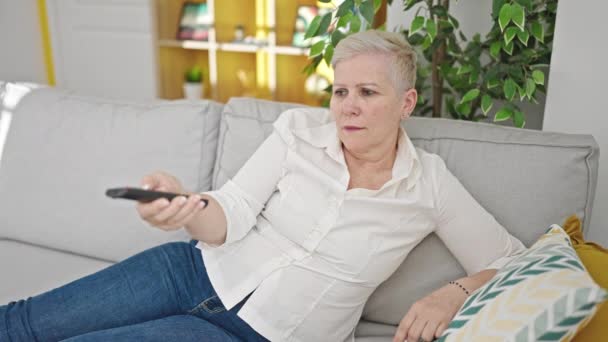 Middle age grey-haired woman watching tv lying on sofa smiling at home - Footage, Video