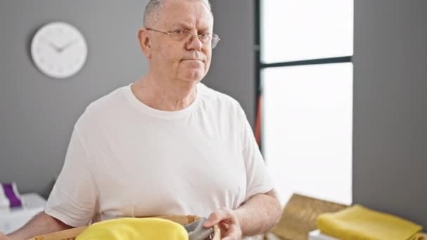 Grauhaariger Mann mittleren Alters lächelt selbstbewusst und hält einen Korb mit Kleidung in der Waschküche - Filmmaterial, Video
