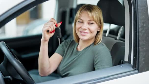 Młoda blondynka uśmiecha się pewnie trzymając klucz nowego samochodu na ulicy - Materiał filmowy, wideo
