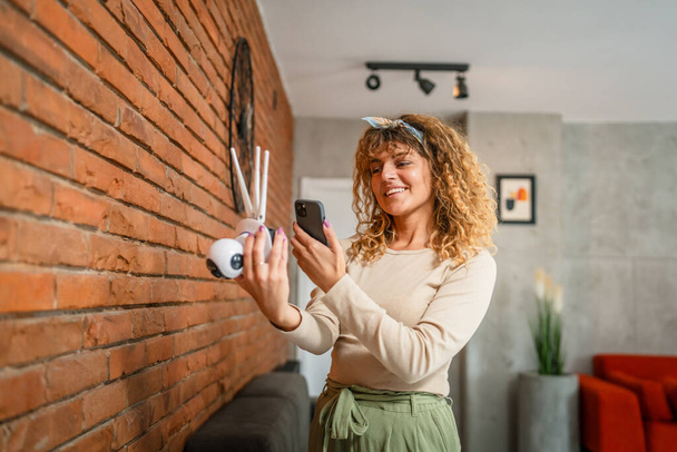 Eine erwachsene kaukasische Frau passt die Anwendung für Überwachungskameras zu Hause an und nutzt das Smartphone, um Kopierraum einzurichten - Foto, Bild