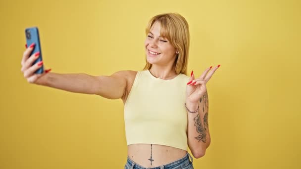 Junge blonde Frau lächelt selbstbewusst und macht Selfie mit dem Smartphone vor einsamem gelben Hintergrund - Filmmaterial, Video