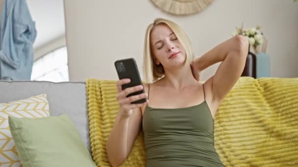 Nuori blondi nainen käyttää älypuhelinta kärsimystä kohdunkaulan kipua kotona - Materiaali, video