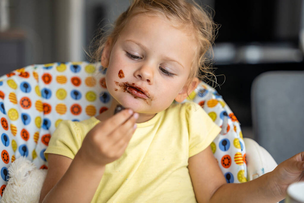 Szőke hajú kislány, házi készítésű csokit eszik piszkos szájjal és kézzel a konyhában. Kiváló minőségű fénykép - Fotó, kép