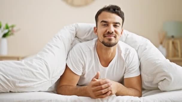 Νεαρός Ισπανός άντρας χαμογελάει με αυτοπεποίθηση ξαπλωμένος στο κρεβάτι στην κρεβατοκάμαρα - Πλάνα, βίντεο