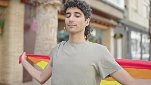 Joven hispano sosteniendo bandera de arco iris con expresión relajada en la calle - Imágenes, Vídeo