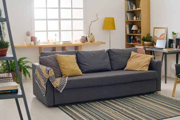 Teil des geräumigen Wohnzimmers mit grauer bequemer Couch mit einer Gruppe von Kissen, die in der Mitte auf dem Boden stehen und mit gestreiftem Teppich bedeckt sind - Foto, Bild