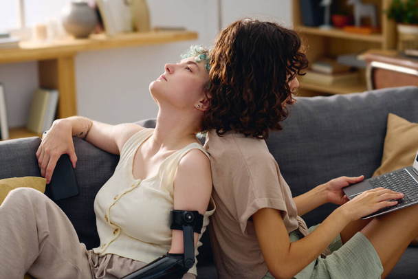 Молода спокійна жінка з міоелектричною рукою сидить на дивані проти своєї дівчини, друкуючи на клавіатурі ноутбука під час спілкування або перегляду відео - Фото, зображення
