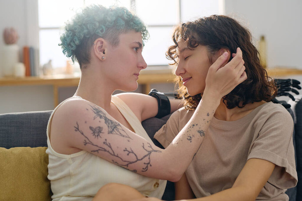 Молодая женщина с миоэлектрической рукой трогает волосы своей безмятежной ласковой подружки, сидящей рядом с ней и наслаждающейся интимным моментом - Фото, изображение