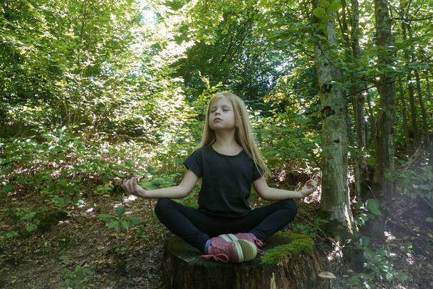 Kleines Mädchen in Lotusposition auf dem Baumstumpf sitzend. Waldmeditation. Ruhe, Achtsamkeit und Entspannung in der Natur. - Foto, Bild