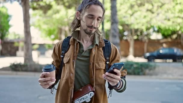 Νεαρός Ισπανόφωνος τουρίστας χρησιμοποιώντας smartphone κρατώντας καφέ στο πάρκο - Πλάνα, βίντεο