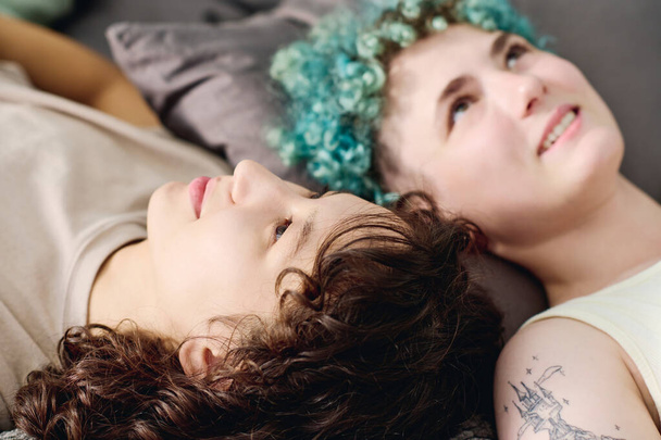 Focus on face of young serene brunette woman lying on bed in front of camera against her smiling girlfriend with short blue curly hair - Фото, зображення