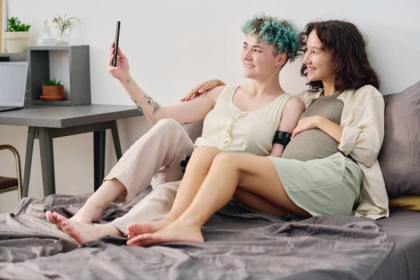 Two happy young girlfriends sitting on double bed and looking at smartphone screen while woman with myoelectric hand taking selfie - Fotoğraf, Görsel