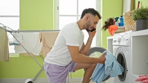 Молодий арабський чоловік миє одяг, розмовляючи по смартфону в пральні - Кадри, відео