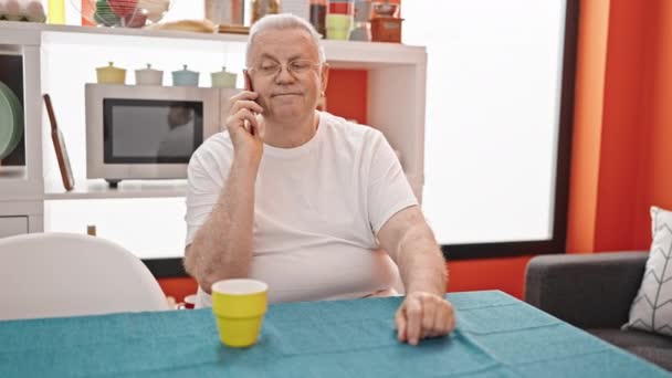 中世の灰色の髪の男性は,ダイニングルームでテーブルに座ってスマートフォンで話しています - 映像、動画