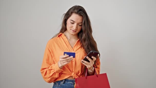 Νεαρή όμορφη ισπανόφωνη γυναίκα που κρατάει τσάντες αγορών χρησιμοποιώντας smartphone και πιστωτική κάρτα σε απομονωμένο λευκό φόντο - Πλάνα, βίντεο