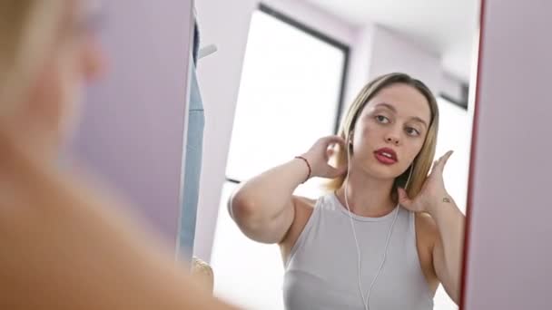 Young blonde woman combing hair with listening to music looking on mirror at home - Footage, Video