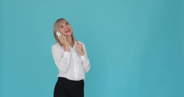 Excited woman is captured talking on the phone with great enthusiasm against a serene blue background. Her animated gestures and lively expression reflect her excitement. - Footage, Video